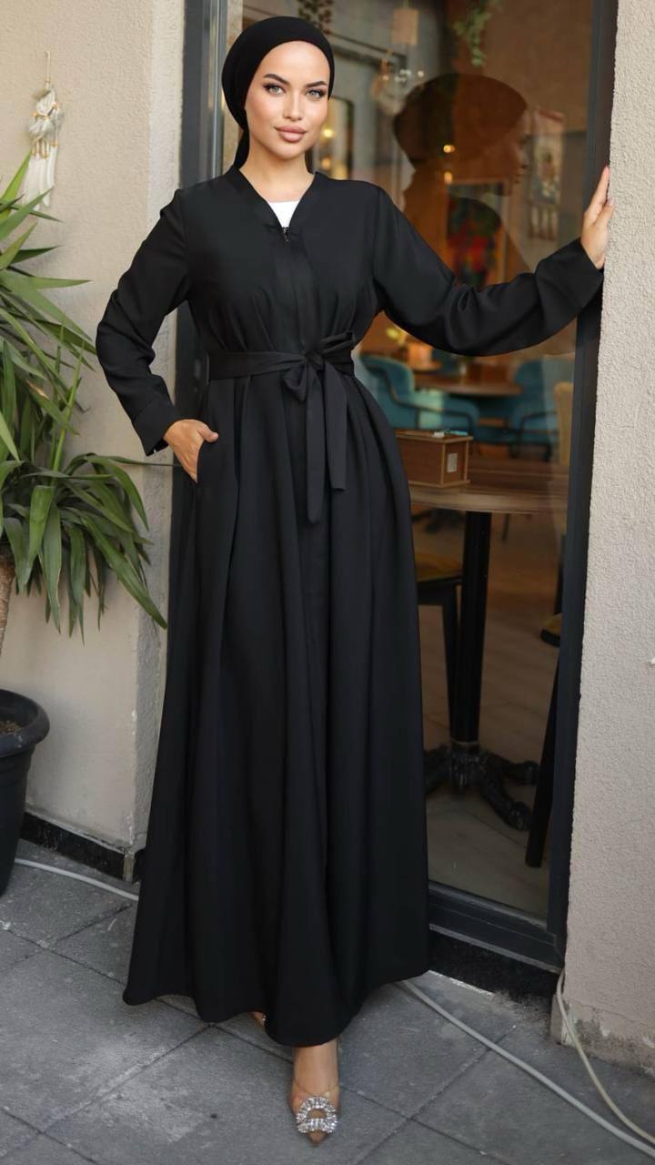 Woman wearing a black zippered abaya with a waist belt, standing in front of a café. The abaya has a flowy design and long sleeves, paired with a matching hijab and embellished heels.