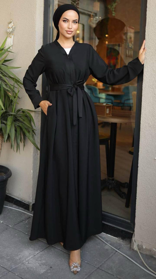 Woman wearing a black zippered abaya with a waist belt, standing in front of a café. The abaya has a flowy design and long sleeves, paired with a matching hijab and embellished heels.