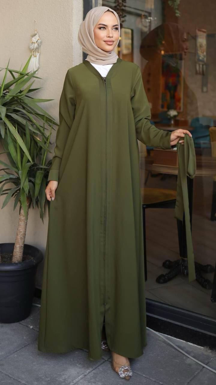 Woman wearing a green zippered abaya with a waist belt, standing in front of a café. The abaya has a flowy design and long sleeves, paired with a matching hijab and embellished heels.