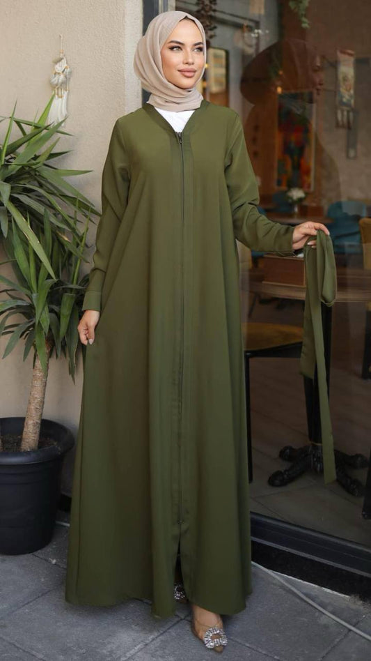 Woman wearing a green zippered abaya with a waist belt, standing in front of a café. The abaya has a flowy design and long sleeves, paired with a matching hijab and embellished heels.