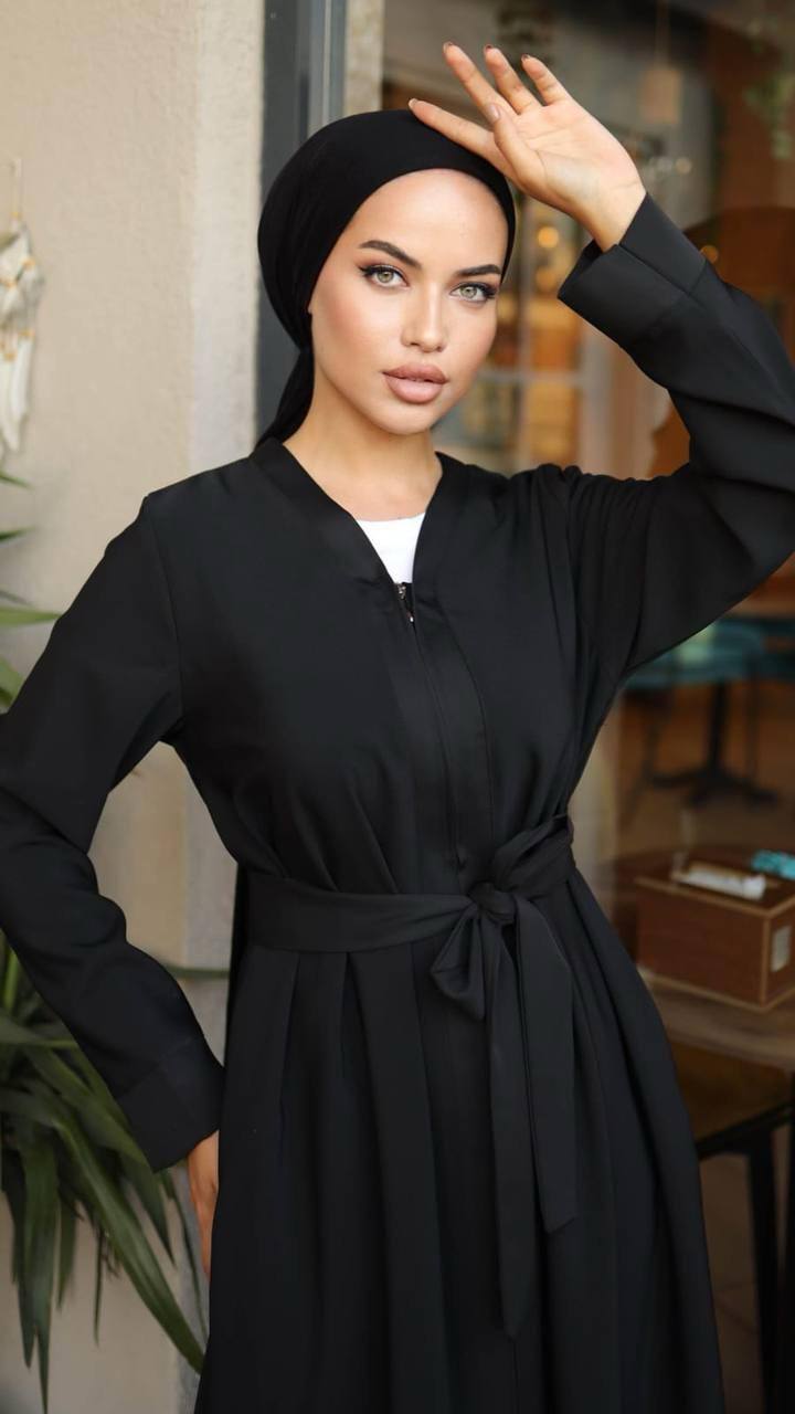 Close-up of Woman wearing a black zippered abaya with a waist belt, standing in front of a café. The abaya has a flowy design and long sleeves, paired with a matching hijab and embellished heels.