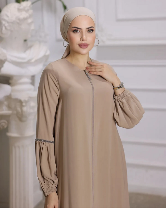 woman wearing a zipper-front abaya with elegant puff sleeves, a flowing silhouette, side pockets, and a modest fit, paired with a matching hijab and embellished flats.