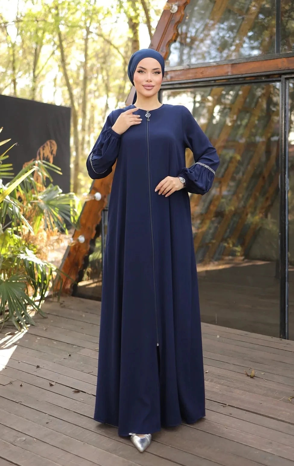 A woman wearing a zipper-front abaya with elegant puff sleeves, a flowing silhouette, side pockets, and a modest fit, paired with a matching hijab and embellished flats.