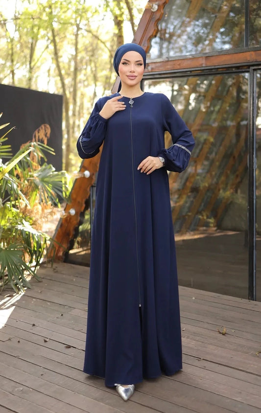 A woman wearing a zipper-front abaya with elegant puff sleeves, a flowing silhouette, side pockets, and a modest fit, paired with a matching hijab and embellished flats.