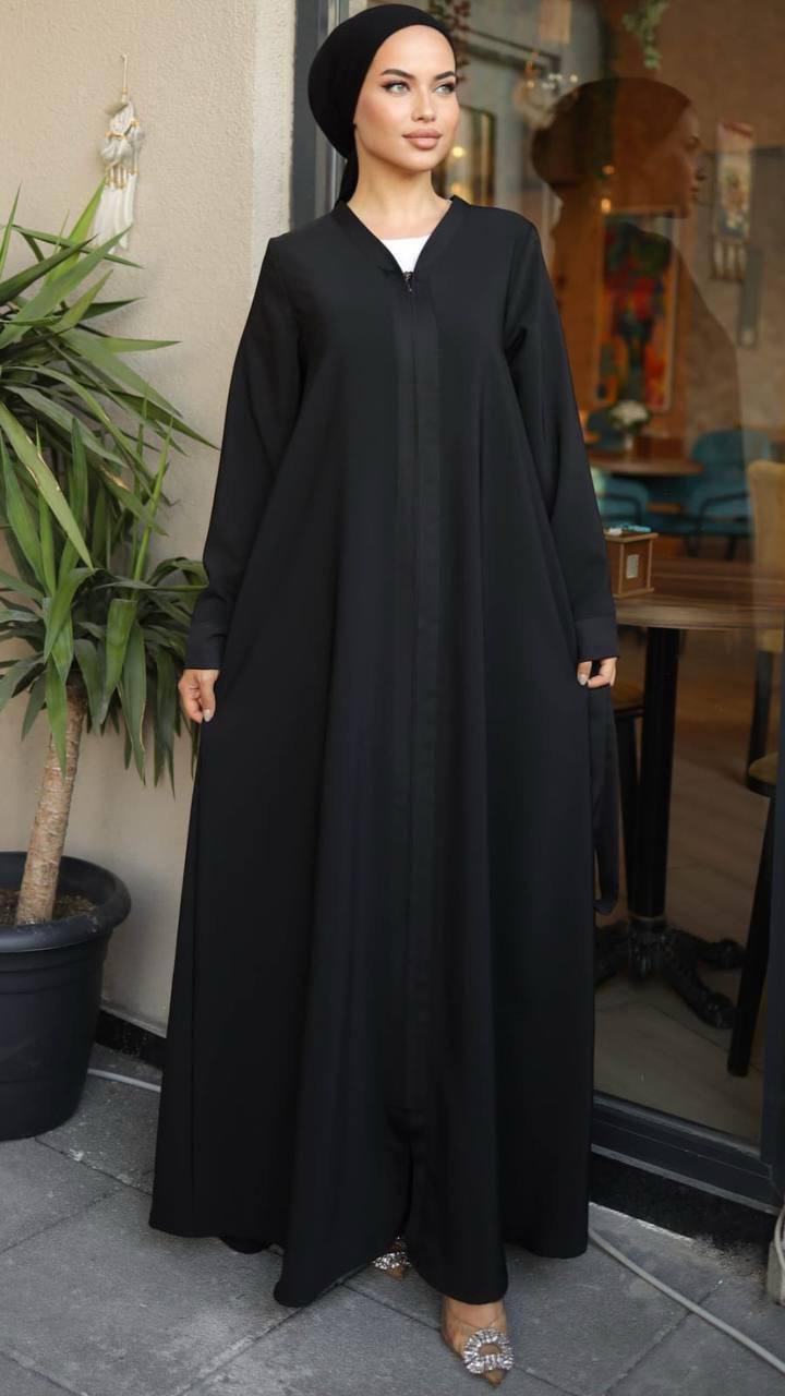 Woman wearing a black flowy zippered abaya. The abaya has a flowy design and long sleeves, paired with a matching hijab and embellished heels.