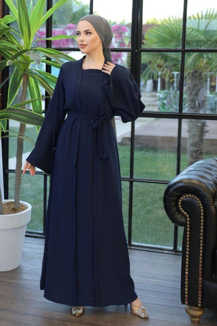 A woman wearing a one-piece kimono-style abaya with a matching belt, long wide sleeves, and a modest silhouette, paired with a hijab and embellished flats.