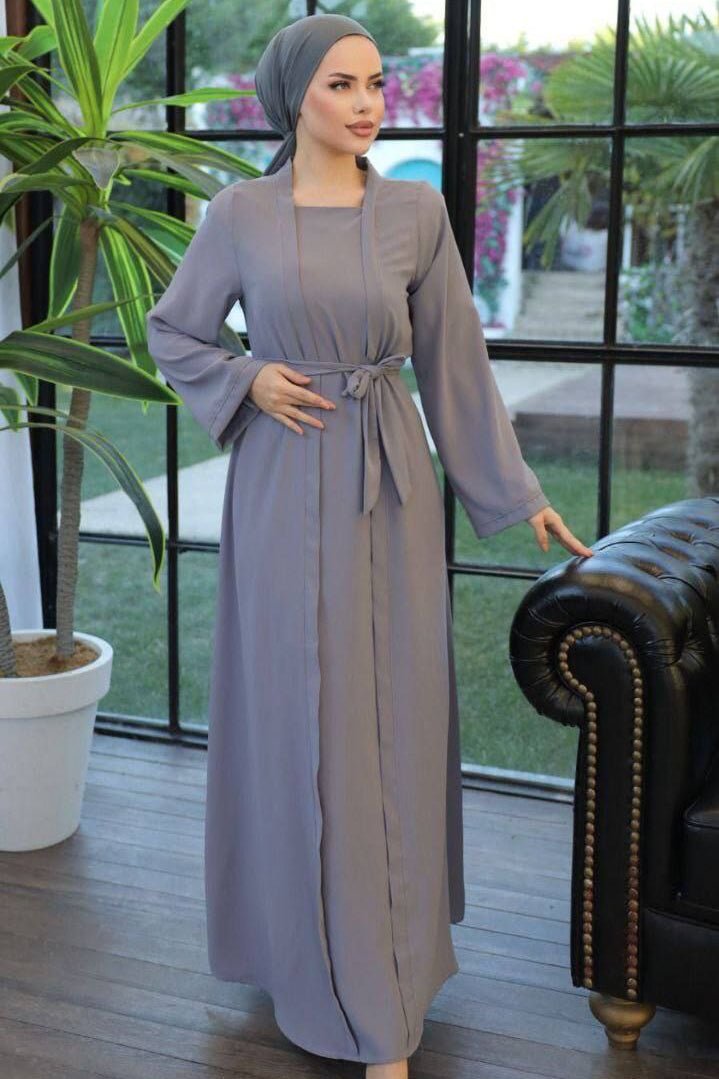 A woman wearing a one-piece kimono-style abaya with a matching belt, long wide sleeves, and a modest silhouette, paired with a hijab and embellished flats.