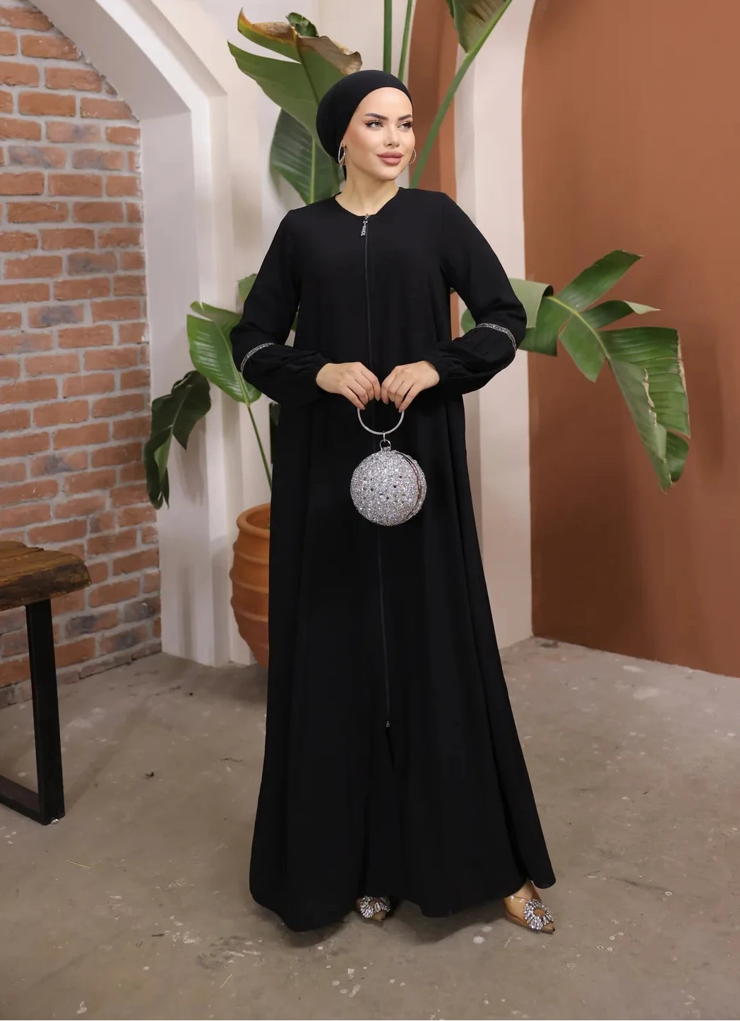 A woman wearing a zipper-front abaya with elegant puff sleeves, a flowing silhouette, side pockets, and a modest fit, paired with a matching hijab and embellished flats.