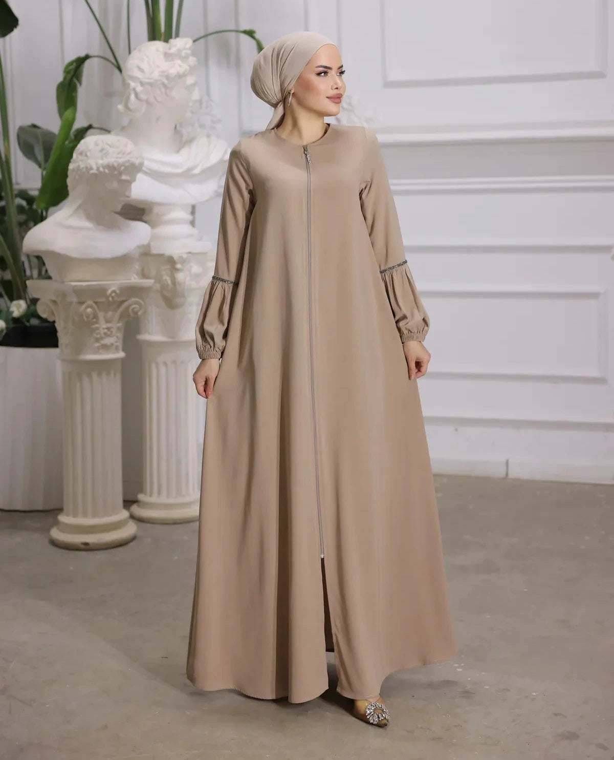 woman wearing a zipper-front abaya with elegant puff sleeves, a flowing silhouette, side pockets, and a modest fit, paired with a matching hijab and embellished flats.