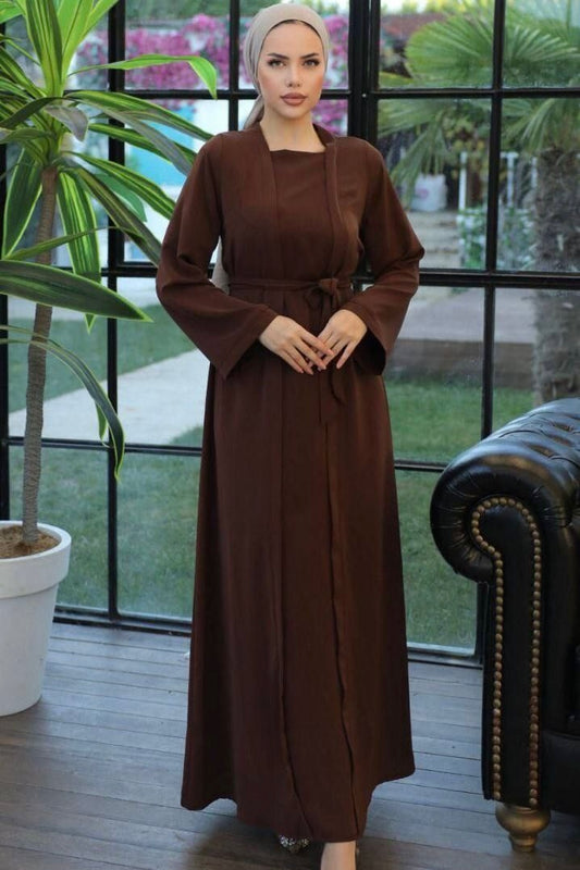  A woman wearing a one-piece kimono-style abaya with a matching belt, long wide sleeves, and a modest silhouette, paired with a hijab and embellished flats.